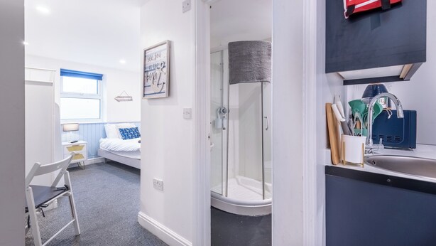 Une chambre d’hôtel compacte comprenant un lit, un petit bureau, une chaise, une douche et une kitchenette équipée d’un évier et d’ustensiles de cuisine.