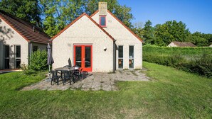 A single-story building with a red door, a small window, and a chimney. There is a patio with a table and chairs. - Ewijk Lake Bungalow With Garden (Ewijk)