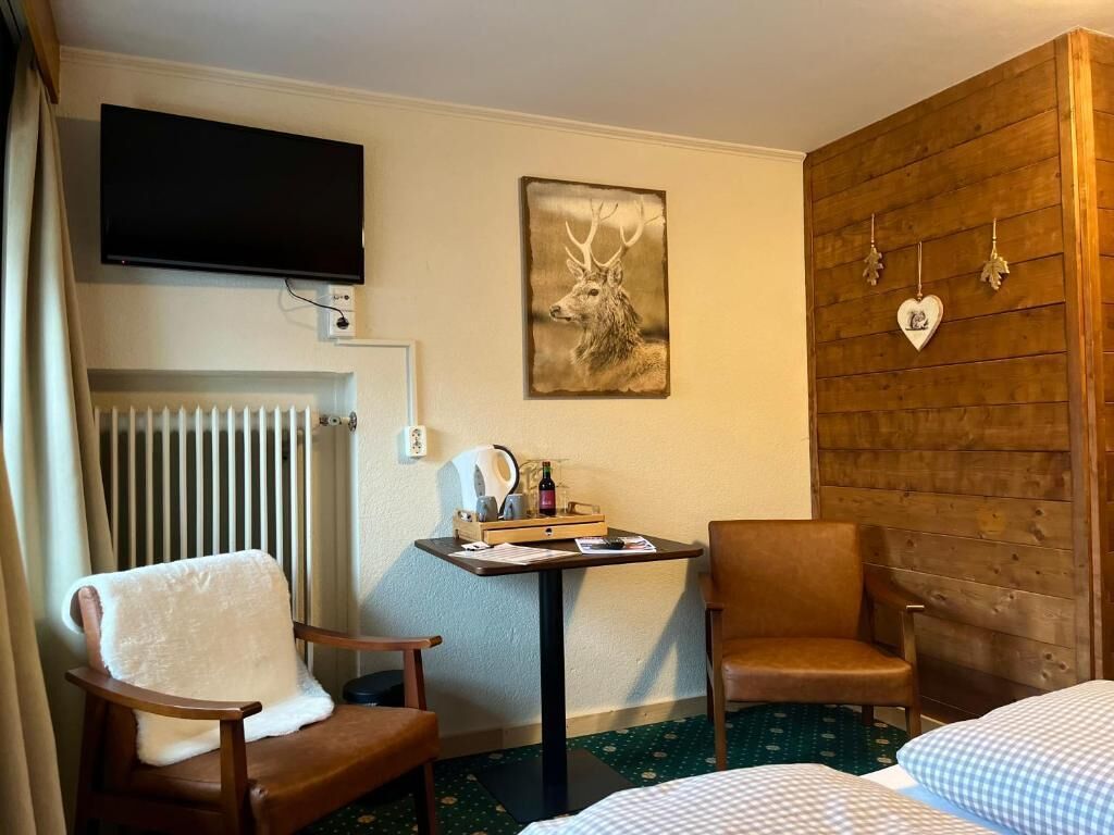 A room with a bed, two chairs, a table with a kettle and a bottle, a television, a deer artwork, and a wooden wall with heart-shaped decorations.