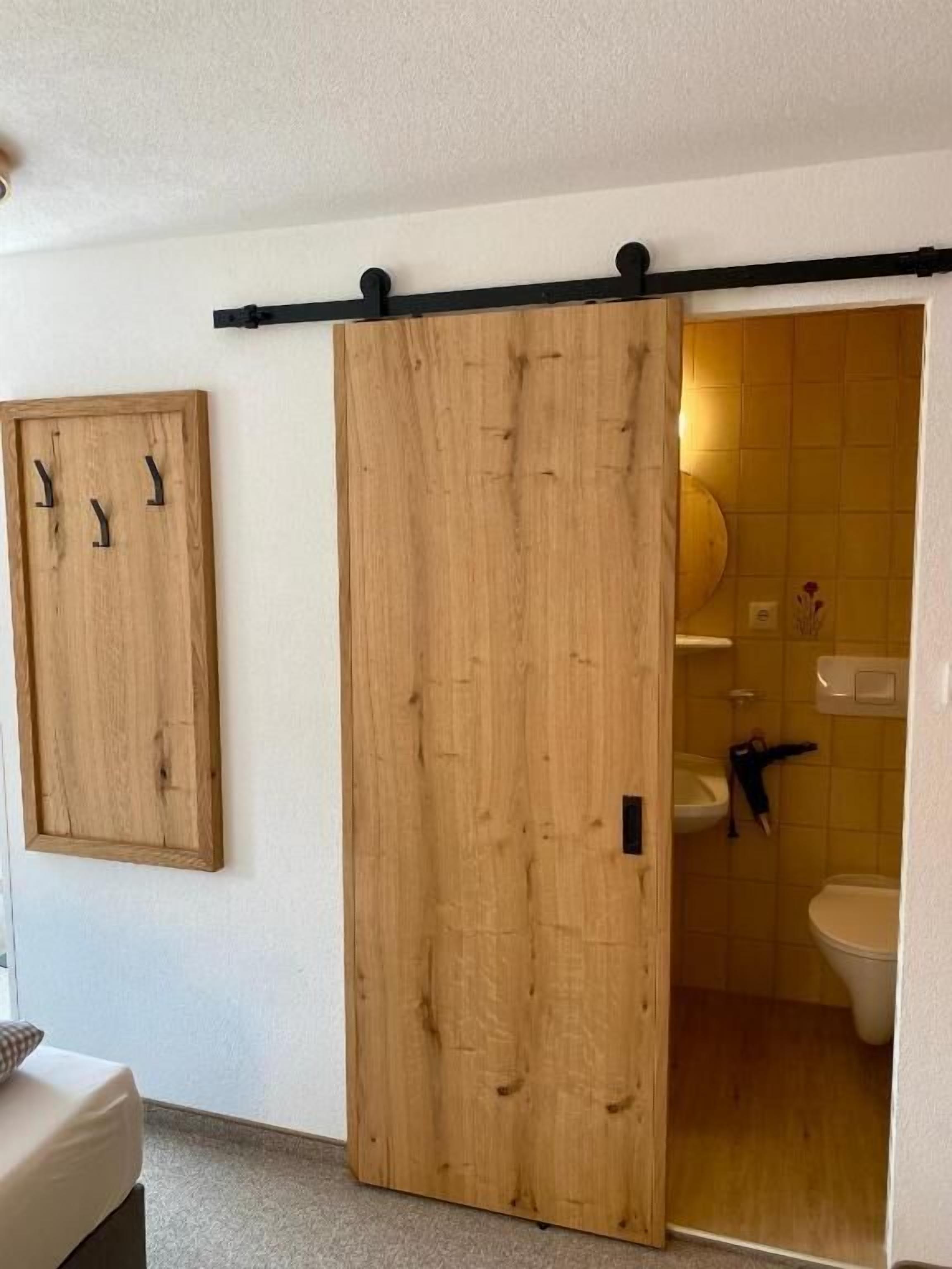 Ein Zimmer mit Holztür zum Badezimmer, einem Holzkabinet mit Haken und einem Bett mit Kissen.