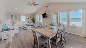 A spacious living area with a dining table, seating, a ceiling fan, and large windows with a view of the ocean. - JL #838 | Deja View | Oceanfront | Ocean Views (Kill Devil Hills)