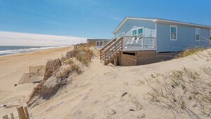 A beachfront house with a wooden staircase leading to a deck, surrounded by sand and sparse vegetation. - JL #838 | Deja View | Oceanfront | Ocean Views (Kill Devil Hills)
