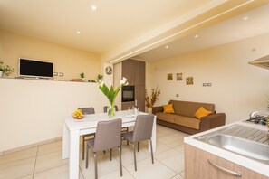 A modern kitchen with a built-in stove, a dining area with a white table and chairs, and a living area with a brown sofa and a TV mounted on the wall.