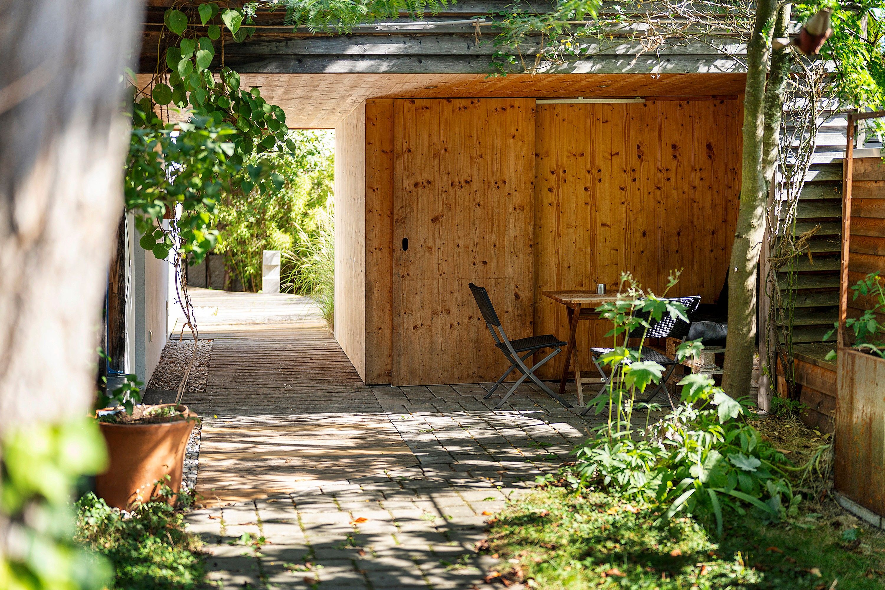 A wooden outdoor seating area with a table and chairs, surrounded by plants and a wooden fence.