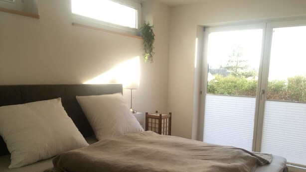 Ein Schlafzimmer mit Bett, Kissen, Nachttisch, Lampe und Blick durch eine Schiebetür.