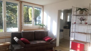 Ein Wohnzimmer mit einer Couch, einem roten Teppich, einem Korbstuhl, einem Metallregal mit Pflanzen und Blick ins Freie durch große Fenster.