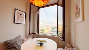 Ein Zimmer mit einem runden Tisch, zwei gemusterten Kissen, einem gerahmten Bild und einem Fenster mit freiem Blick auf eine Stadtlandschaft.