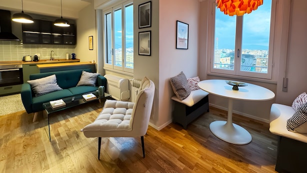 Ein modernes Wohnzimmer mit Sofa, Sessel, rundem Tisch und Blick auf die Stadt durch große Fenster.