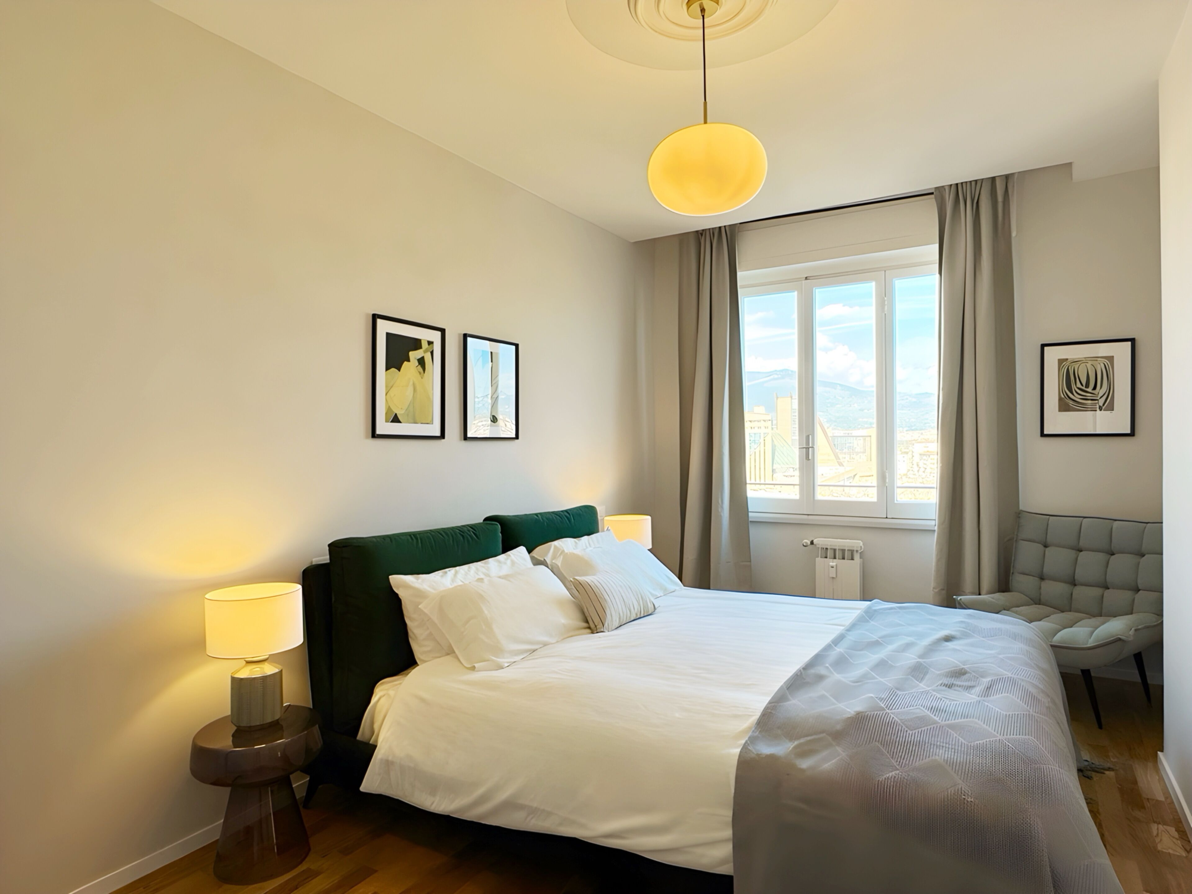 A hotel room with a bed, two bedside lamps, a window with curtains, a chair, and two framed pictures on the wall.