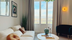 Ein modernes Wohnzimmer mit einer Couch, einem runden Couchtisch, einem hängenden, grünen Licht und Blick durch ein großes Fenster.