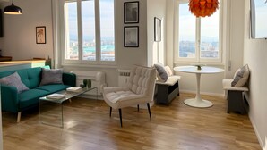 Ein modernes Wohnzimmer mit Sofa, Sessel und Couchtisch, großen Fenstern mit Blick auf die Stadt und einer auffälligen, orangefarbenen Pendelleuchte.