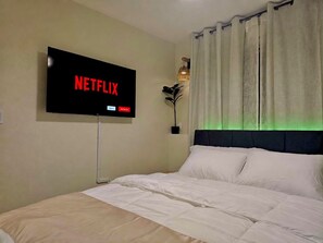 Habitación de hotel con cama, televisión instalada en la pared mostrando el logo de Netflix, planta en maceta y ventana con cortinas.