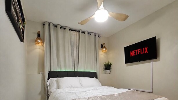 Un dormitori amb un llit, un ventilador de sostre, un televisor fixat a la paret que mostra 'NETFLIX' i una petita planta en test.