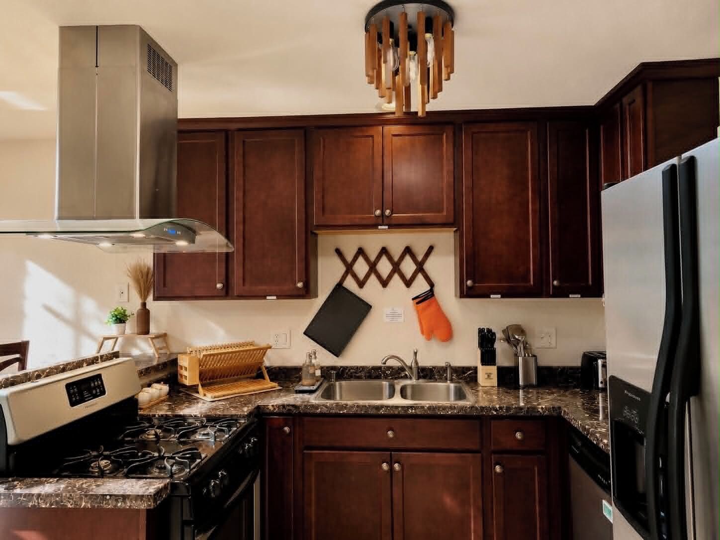 Une cuisine avec des meubles en bois foncé, une hotte de cuisinière en acier inoxydable, un plan de travail en granit et un luminaire suspendu moderne.