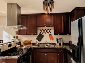 Una cocina con alacenas de madera oscura, una campana extractora de acero inoxidable, una encimera de granito y una lámpara colgante moderna.