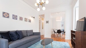 Olohuoneessa on harmaa sohva, sohvapöytä ja TV-taso. Seinällä on koristeellisia laattoja, ja taustalla näkyy ruokailutila.