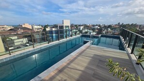 A rooftop swimming pool with glass railings, a wooden deck, and a cityscape view in the background. - Apt a 120m das piscinas naturais by LP (Ipojuca)