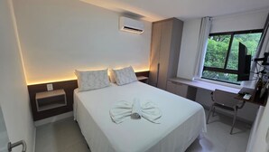 Ein Hotelzimmer mit Bett, Schreibtisch, Stuhl und einem Fenster mit Blick auf Grünflächen, sowie einer Klimaanlage.