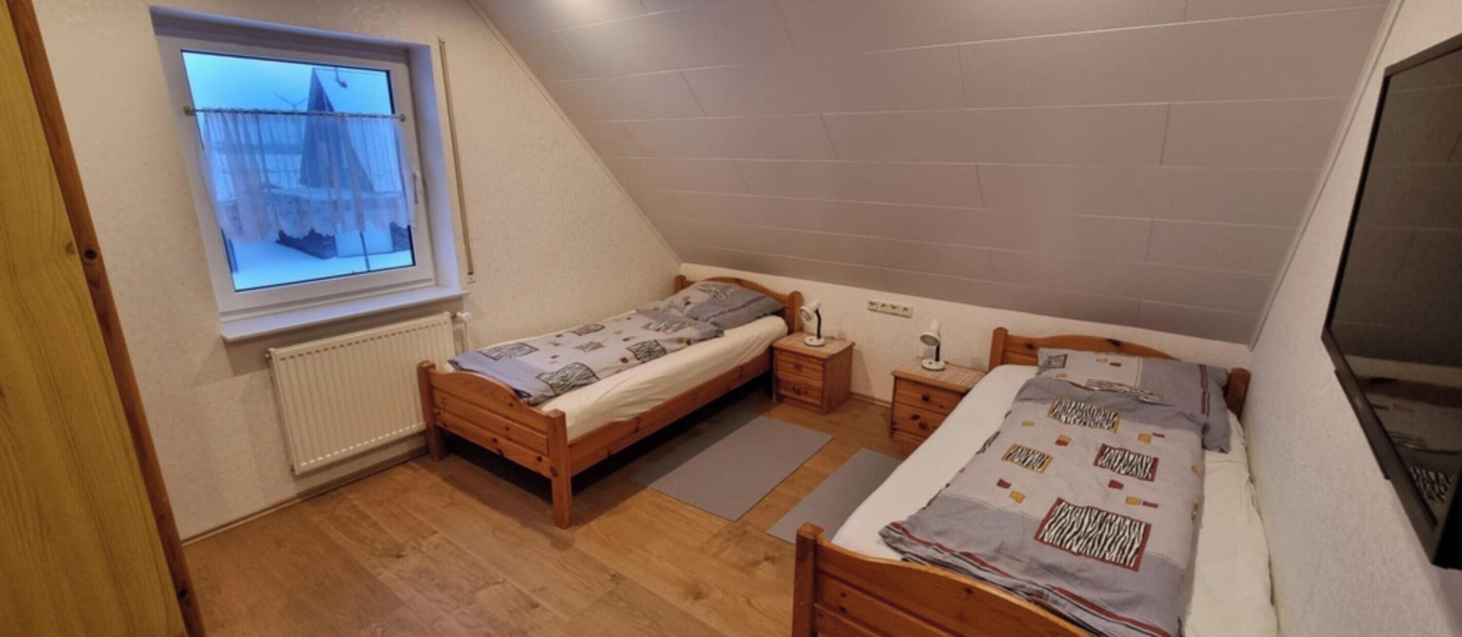 Una habitación pequeña de un dormitorio con techo inclinado, ventana con vistas, dos mesitas de noche y una cama con colcha estampada.