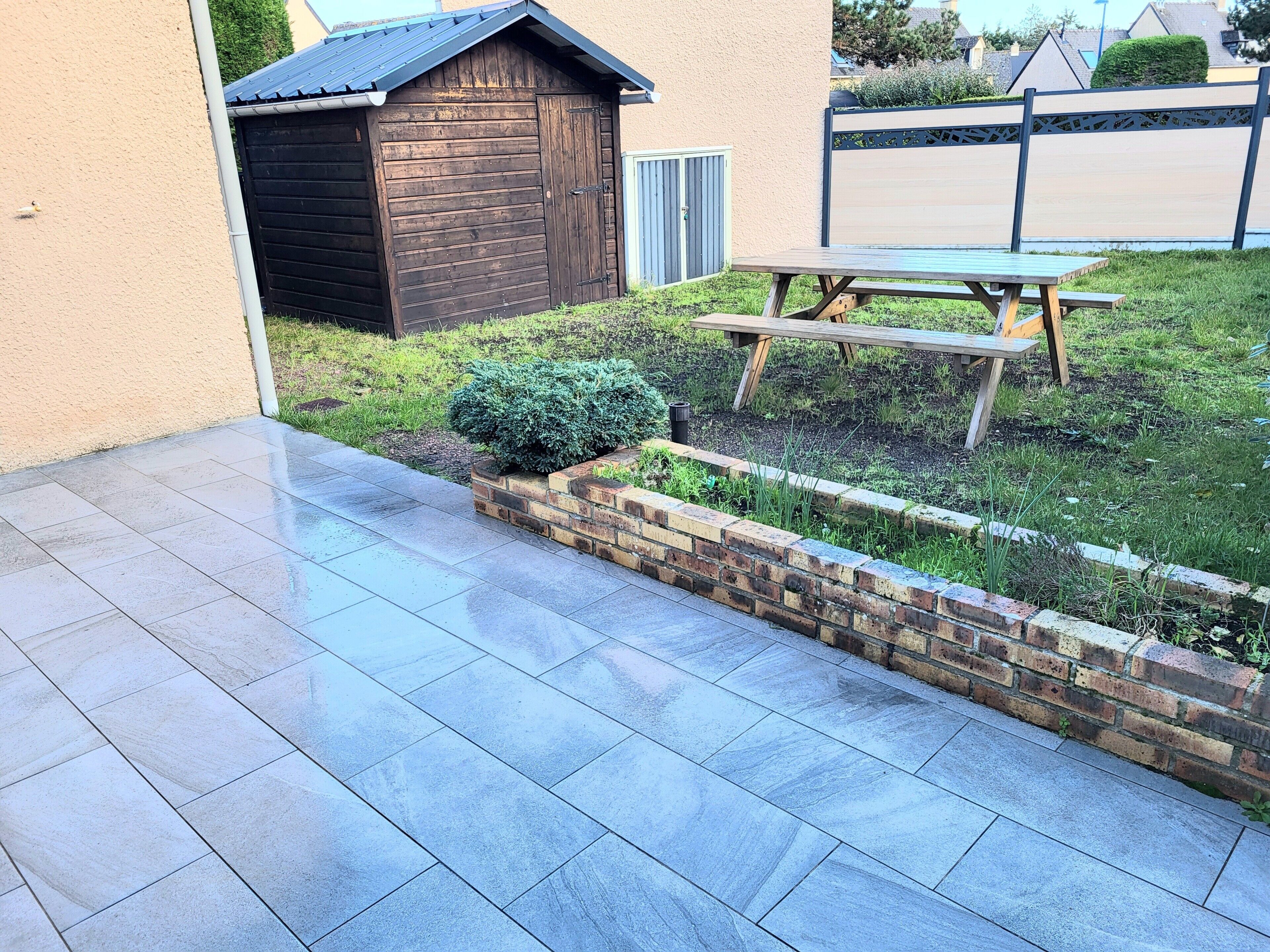 A patio with a tiled floor, a wooden shed, a wooden picnic table, and a brick planter with greenery.