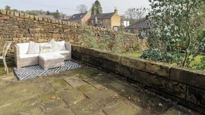 Un muro de piedra, un sofá de mimbre con cojines y una alfombra estampada en un patio de piedra.