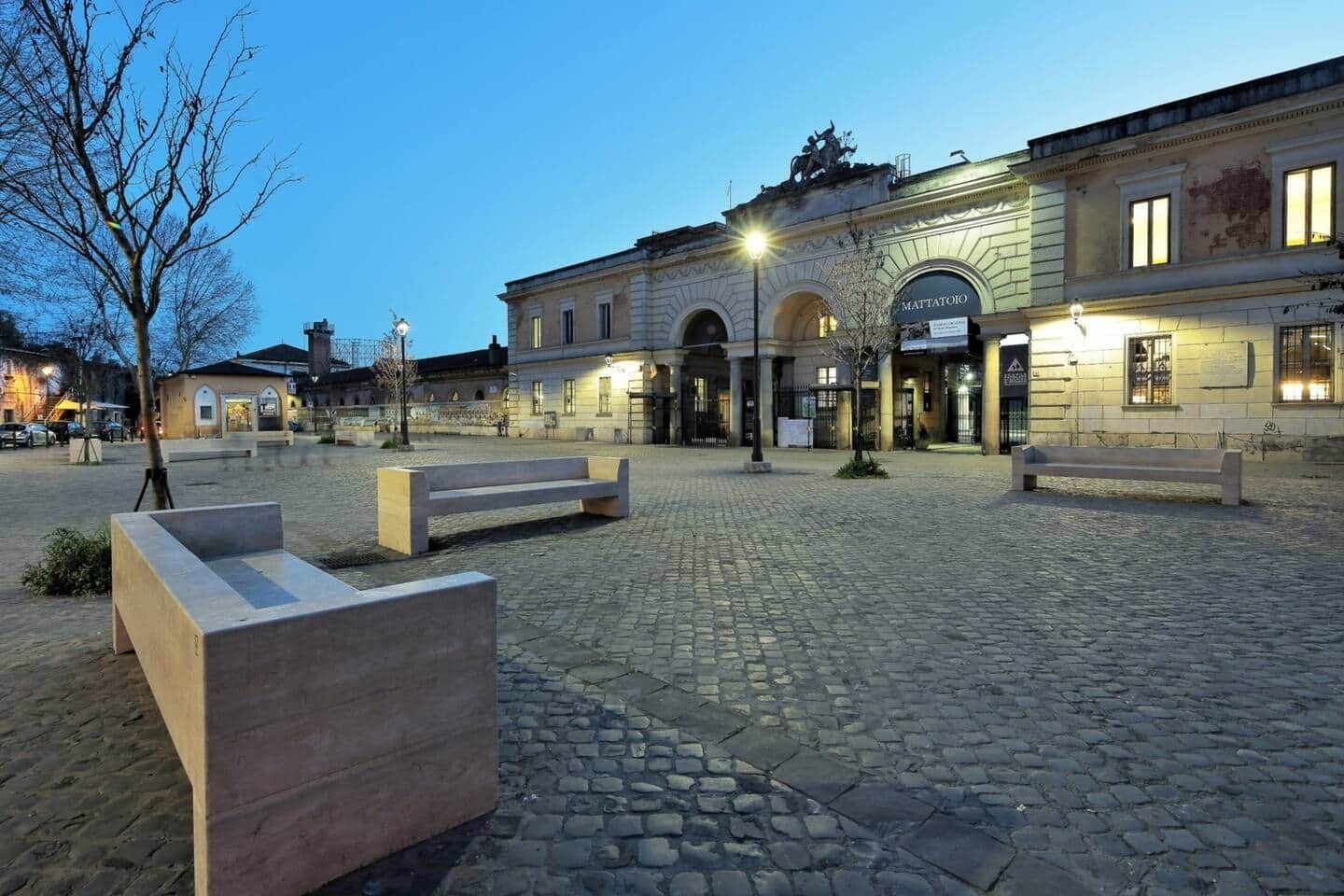 A cobblestone plaza with benches, a building with arched entrances, and a statue on top.