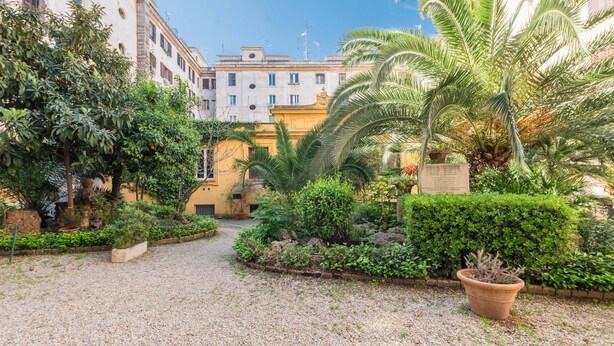 A courtyard with a gravel path, lush greenery, and a building with multiple windows and balconies. - Marmorata 169 - Loft in Testaccio (Rome)