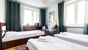 A hotel room with two single beds, a desk, a chair, a small table, a window with curtains, and a radiator.