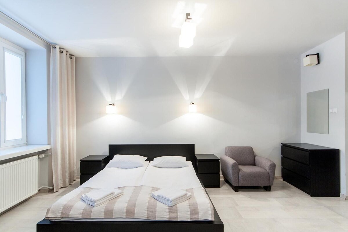 A modern hotel room with a bed, two bedside tables, a chair, a dresser, and a wall-mounted TV.