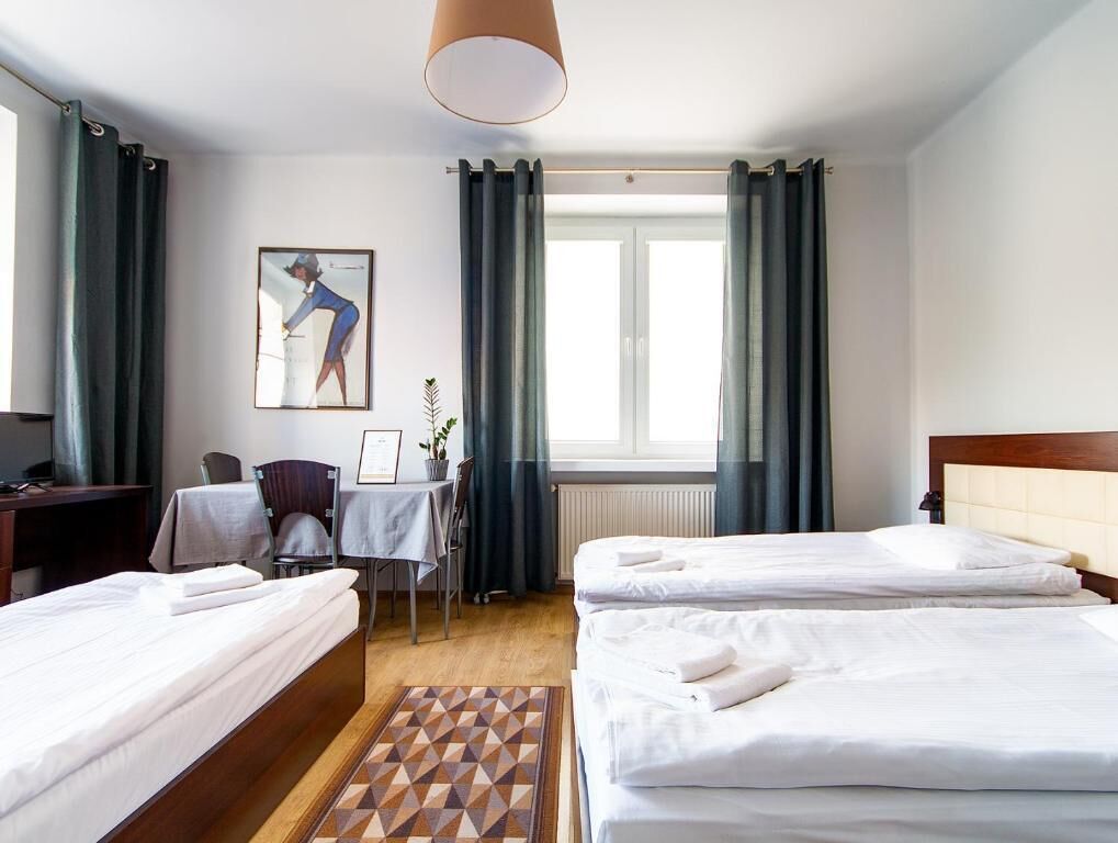 A hotel room with two beds, a desk, a chair, a table, a radiator, and a window with curtains.