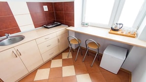 A compact kitchen with a sink, two chairs, a small refrigerator, and a window with a kettle and cups on the counter.