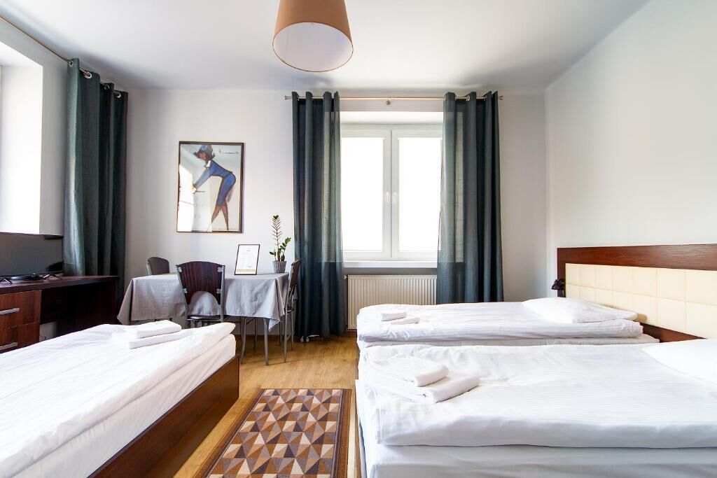 A hotel room with two beds, a desk, a chair, a table, a window with curtains, and a painting on the wall.