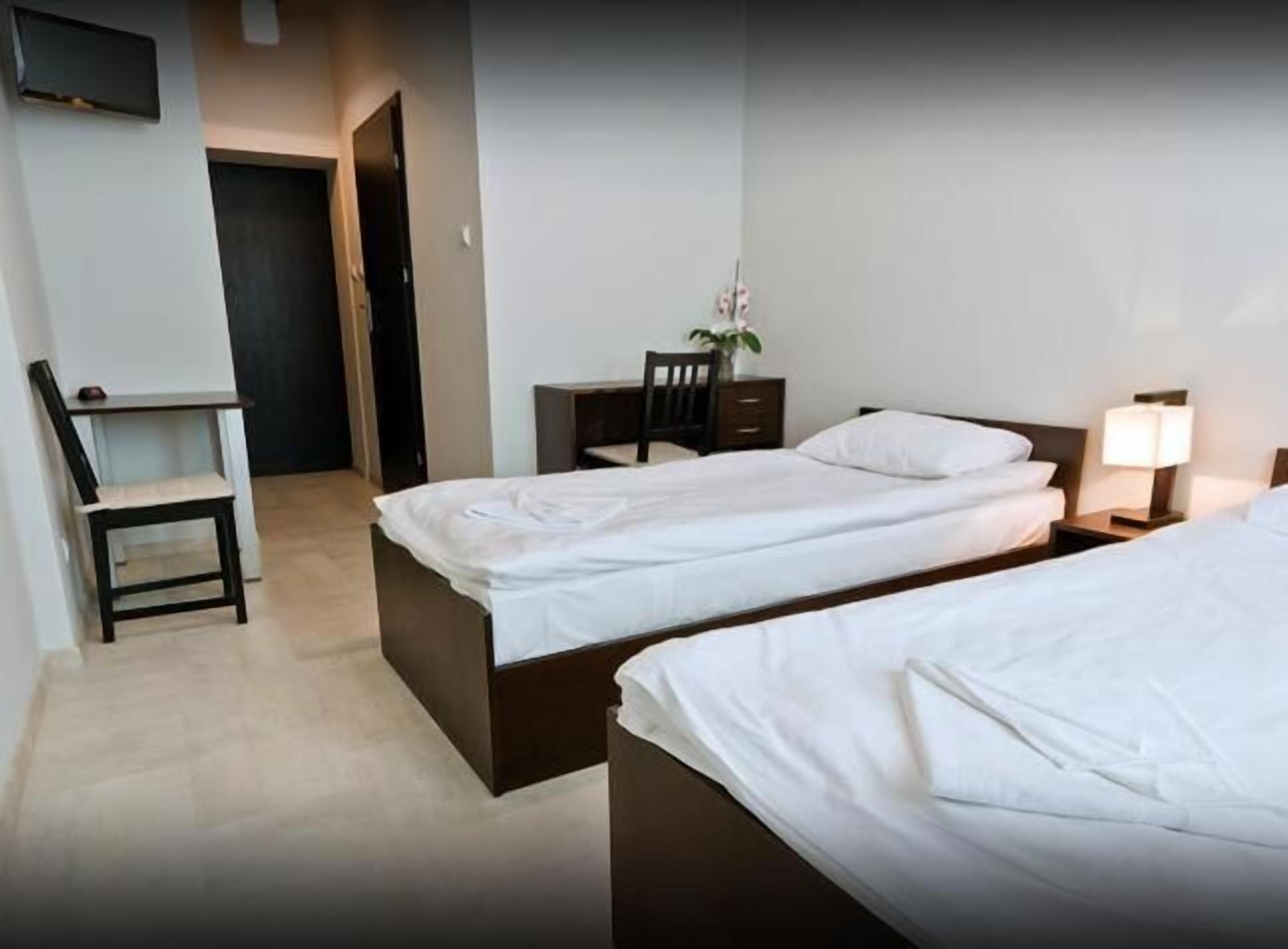 A hotel room with two beds, a desk, a chair, a nightstand with a lamp, and a small table with a vase of flowers.
