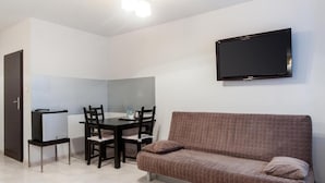 A room with a brown sofa, a dining table with chairs, a flat-screen TV mounted on the wall, and a door.