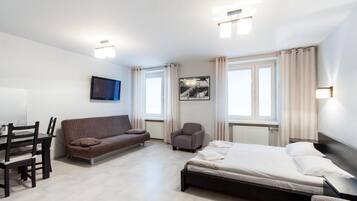 A modern hotel room with a bed, a sofa, a chair, a TV, and a window with curtains.