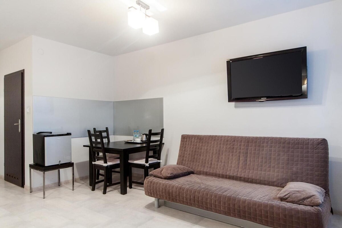 Een compacte kamer met een bruine bank, een eettafel met stoelen, een aan de muur bevestigde televisie en een deur.