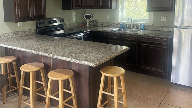 A kitchen with dark wooden cabinets, a granite countertop, a stainless steel refrigerator, and wooden bar stools.