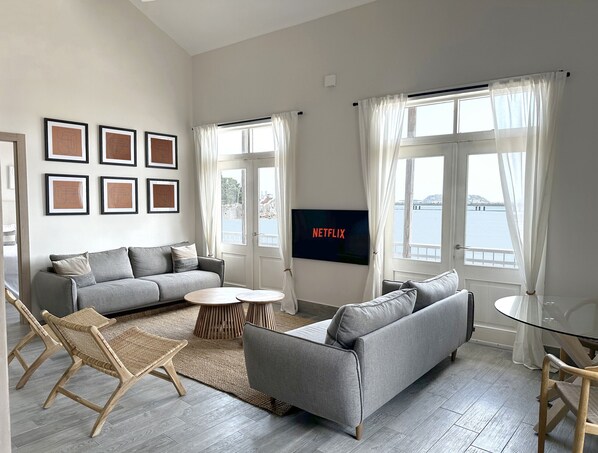 A modern living room with a grey sofa, a small round coffee table, wicker chairs, a flat-screen TV, and a view of the water through large windows. - Unique house with direct access to the beach, four bedrooms, balcony and design (Ciudad de Panamá)