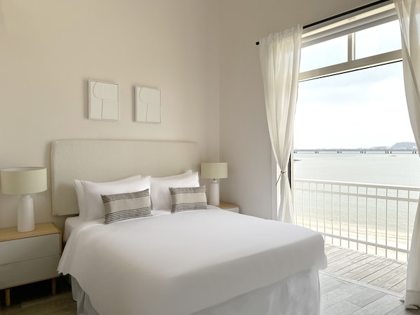 Une chambre d’hôtel avec un grand lit, deux tables de chevet avec des lampes, une vue sur la mer et un balcon.