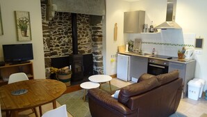 A compact living space with a stone wall, a fireplace, a kitchenette, a brown leather sofa, a dining table, and a television. - Gîte Chèvrefeuille - Pen Ar Voas: Private Terrace, Shared Garden & Wi-Fi (Plestin-les-Grèves)