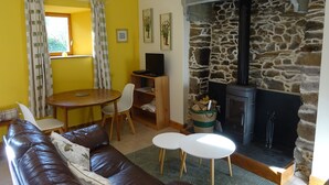 A living room with a stone fireplace, yellow walls, a brown leather sofa, a dining table, and a TV. - Gîte Chèvrefeuille - Pen Ar Voas: Private Terrace, Shared Garden & Wi-Fi (Plestin-les-Grèves)