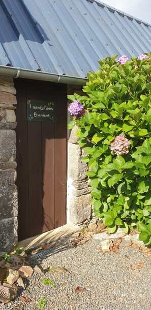 Un edificio de piedra con una puerta de madera que indica 'Laundry Room Byonderie', rodeado de follaje verde y flores moradas.