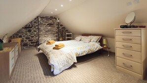 A bedroom with a sloped ceiling, a stone wall, a bed with a patterned bedspread, a dresser with drawers, a bedside lamp, and a mirror on the dresser. - Gîte Chèvrefeuille - Pen Ar Voas: Private Terrace, Shared Garden & Wi-Fi (Plestin-les-Grèves)