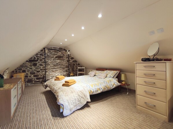 A bedroom with a sloped ceiling, a stone wall, a bed with a patterned bedspread, a dresser with drawers, a bedside lamp, and a mirror on the dresser. - Gîte Chèvrefeuille - Pen Ar Voas: Private Terrace, Shared Garden & Wi-Fi (Plestin-les-Grèves)