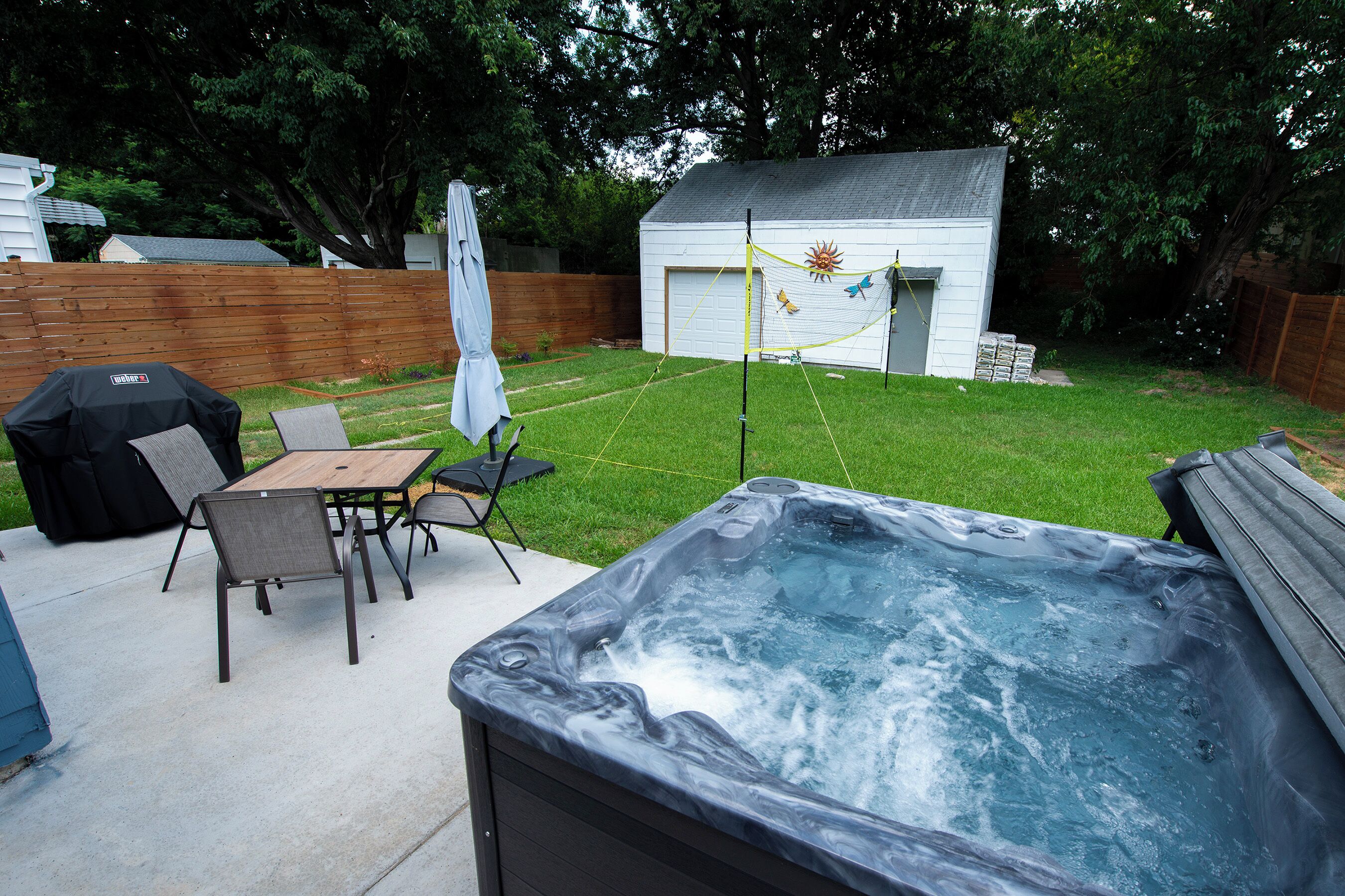 A backyard with a hot tub, a wooden table with chairs, a barbecue grill, and a shed.