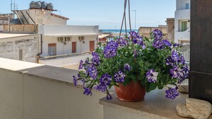 Ein hängender Blumentopf mit violetten Blüten auf einem Balkon mit Blick auf eine Küstenlandschaft und Gebäude.