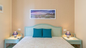Ein Bett mit gemustertem Überwurf, zwei Nachttische mit Lampen, ein türkisblaues Kopfteil und ein gerahmtes Bild mit einer Küstenlandschaft an der Wand.