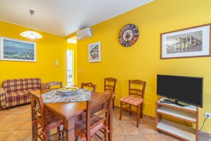 A dining area with a table, chairs, a sofa, a clock, and a television. - ‘Saturno’ Apartment Steps from the Sea with Balcony, Wi-Fi & Air Conditioning (Portopalo di Capo Passero)