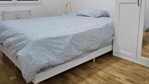 A single-bed room with a patterned bedspread, a radiator, a white wardrobe, and a window with blinds. - Chambre Lit double (Dunkirk)
