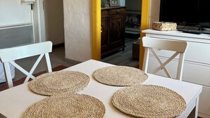 A dining area with a white table, four woven placemats, white chairs, a television, and a kitchen visible in the background. - 5-minute walk from the town center, magnificent view of the sea and the old town (Sète)